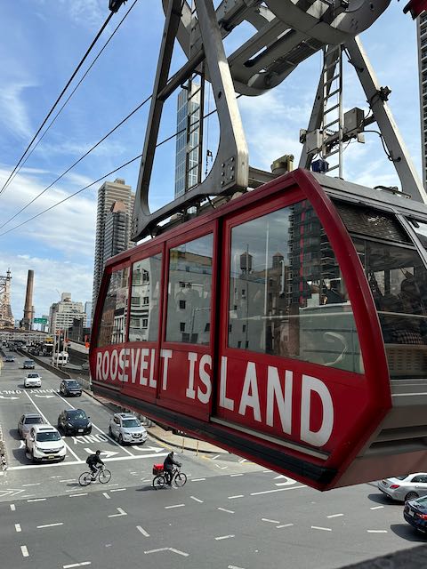 Red cable car suspended above the street with the words Roosevelt Island on it in white text